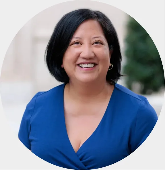 A woman with short black hair, wearing a blue V-neck top, smiles at the camera. She is standing outdoors with a blurred background of a building and greenery.