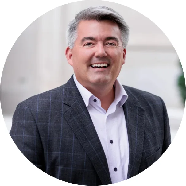 A man with gray hair smiles while wearing a gray plaid blazer and white shirt, standing outdoors in front of a light-colored blurred background.