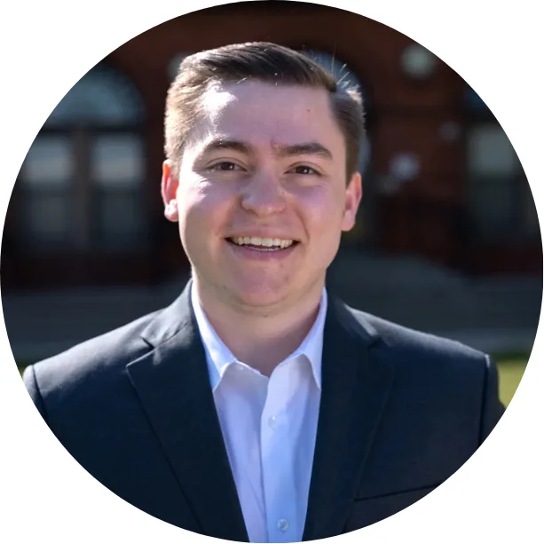 A young man in a dark suit jacket and white shirt smiles at the camera outdoors, with a blurred red brick building and arched windows in the background.