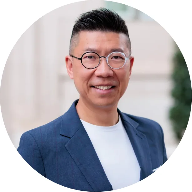 A smiling man with short dark hair and glasses, wearing a blue blazer over a white shirt, stands in front of a blurred light background.