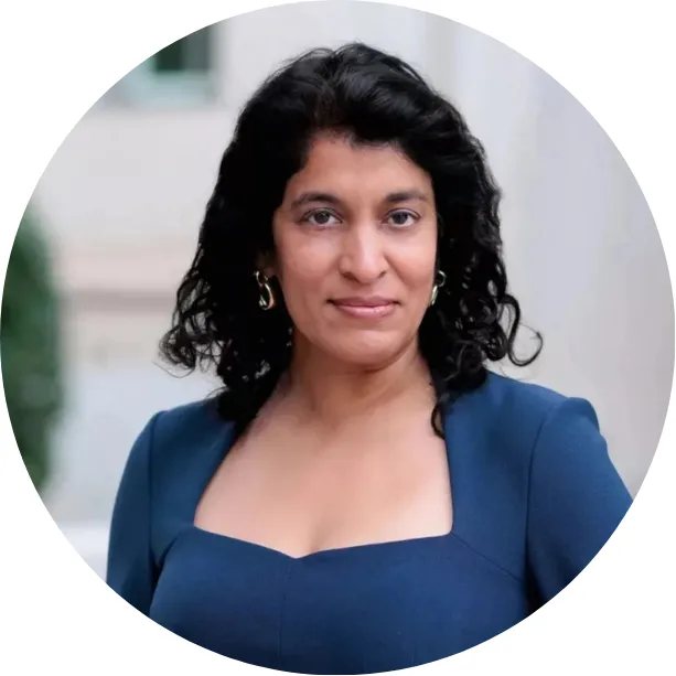 A woman with wavy dark hair, wearing a blue square-neck dress and gold hoop earrings, stands outdoors in front of a blurred light-colored building. She faces the camera with a slight, confident smile.