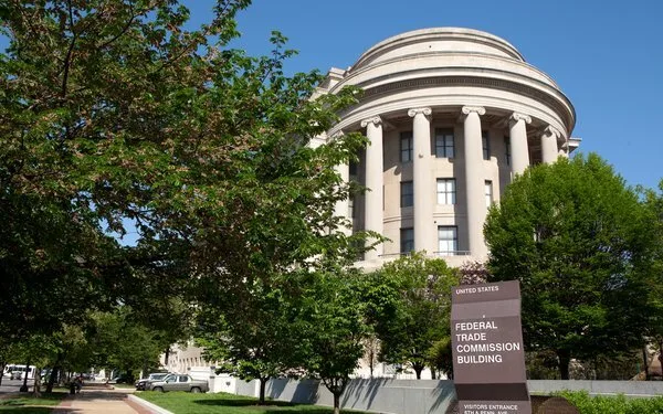 A large, round, neoclassical stone building with columns is surrounded by trees. In front is a sign reading Federal Trade Commission Building with directions to the visitor entrance. The sky is clear and blue.