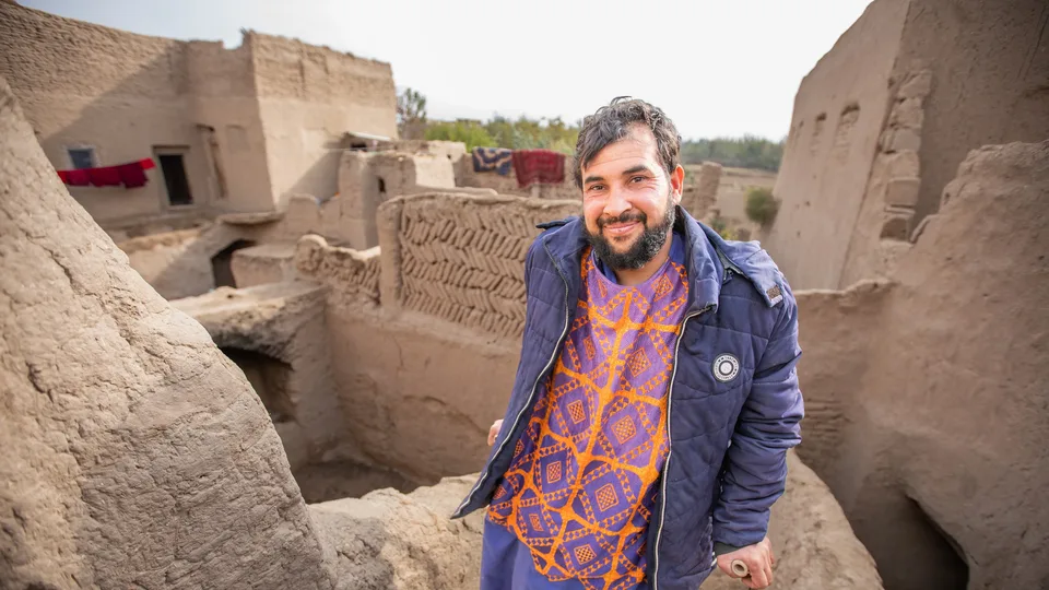 A man wearing a blue jacket and orange patterned shirt stands smiling among mud-brick buildings and walls in a rustic village setting, with rugs hanging in the background.