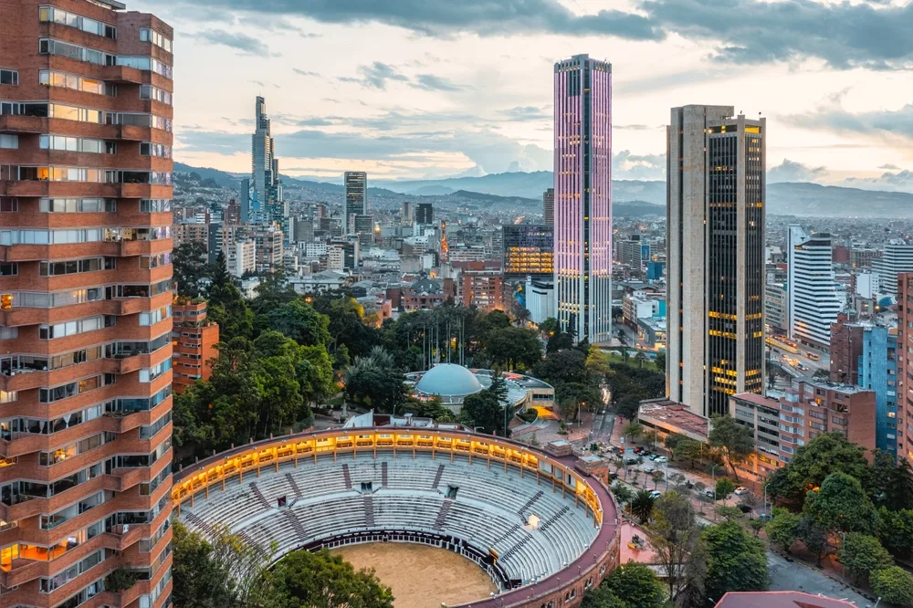 Aerial view of a city with modern skyscrapers and apartment buildings surrounding a large circular bullring, with mountains visible in the background under a cloudy sky.