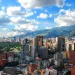 A vibrant cityscape with numerous high-rise buildings, green areas, and mountains in the background under a blue sky with fluffy clouds.
