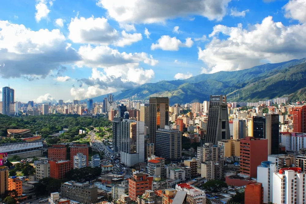 A vibrant cityscape with numerous high-rise buildings, green areas, and mountains in the background under a blue sky with fluffy clouds.