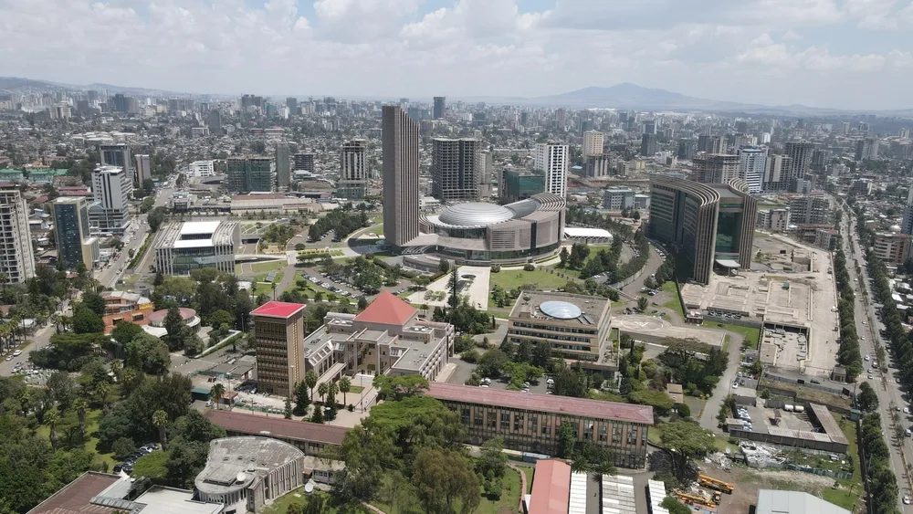 Aerial view of a modern city with tall buildings, greenery, and roads; a round central building and several high-rises dominate the skyline under a partly cloudy sky.