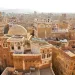 A view of an old city filled with traditional brown brick buildings, domes, and minarets, featuring intricate white detailing, with mountains faintly visible in the hazy background.