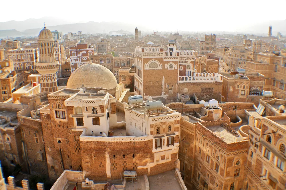 A view of an old city filled with traditional brown brick buildings, domes, and minarets, featuring intricate white detailing, with mountains faintly visible in the hazy background.