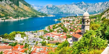 A scenic view of Kotor, Montenegro, showing red-roofed buildings, a stone church tower, lush greenery, and a blue bay surrounded by mountains under a clear sky.