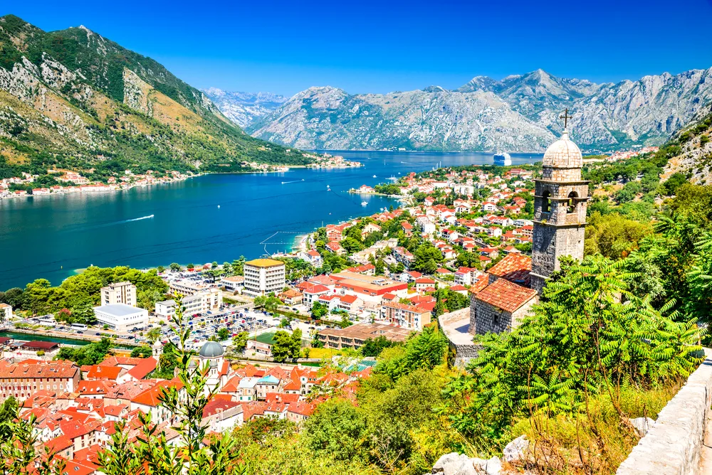 A scenic view of Kotor, Montenegro, showing red-roofed buildings, a stone church tower, lush greenery, and a blue bay surrounded by mountains under a clear sky.