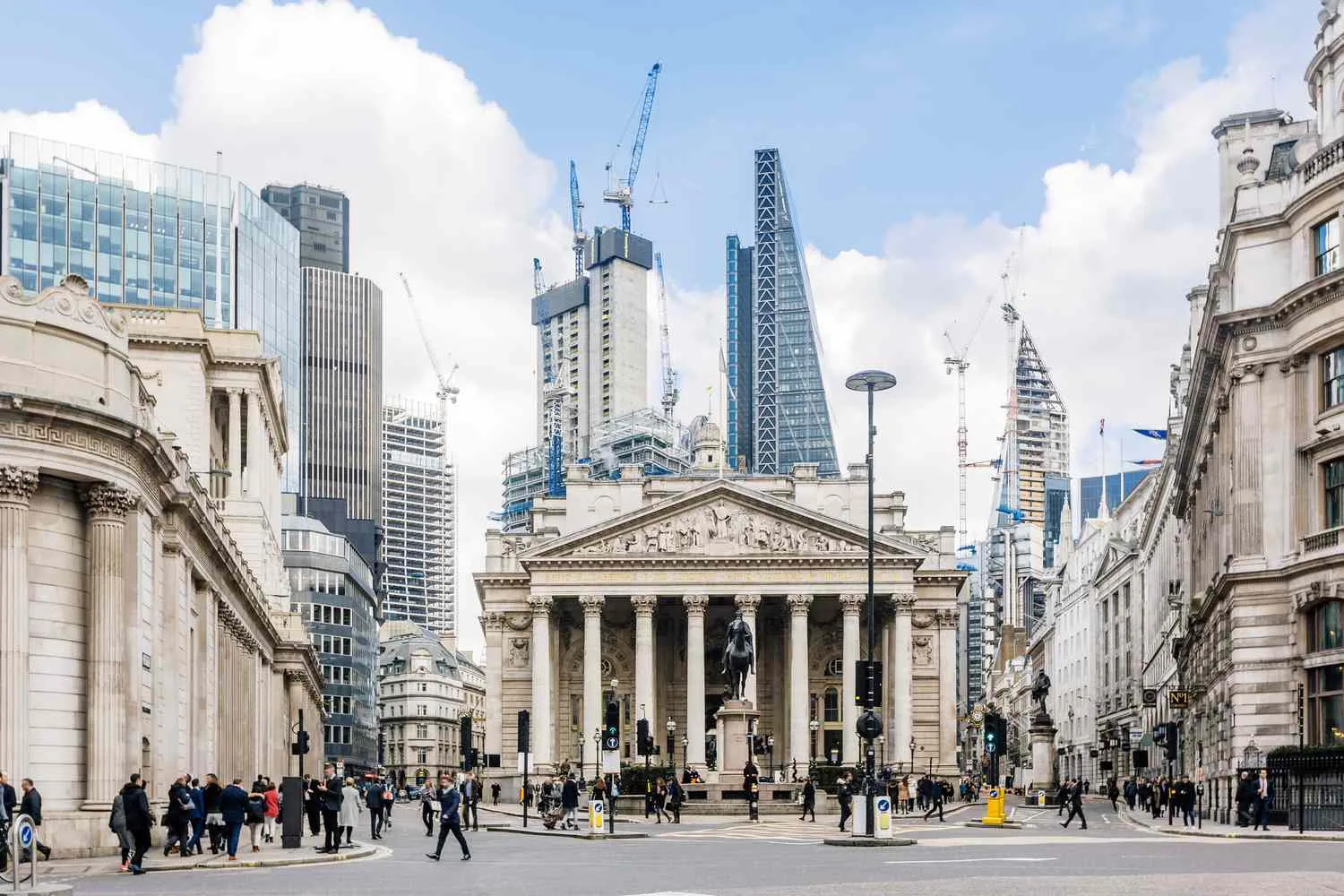 People walk in front of the Bank of England and Royal Exchange in London, surrounded by modern skyscrapers and construction cranes under a partly cloudy sky.